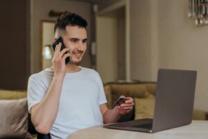 Un jeune homme sourit en faisant des achats en ligne à l'aide d'un téléphone et d'un ordinateur portable, tenant une carte de crédit.